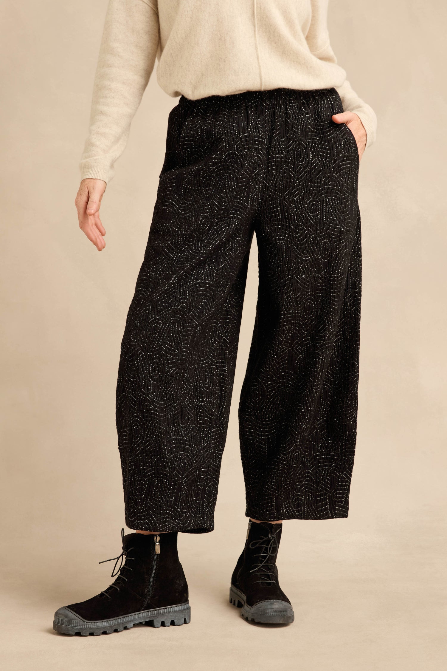 Wearing a cream sweater, the person stands with one hand in a pocket, styled in black Stitched Jacquard Bubble Trousers and black lace-up boots.