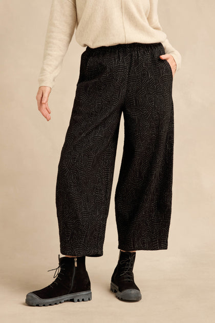 Wearing a cream sweater, the person stands with one hand in a pocket, styled in black Stitched Jacquard Bubble Trousers and black lace-up boots.