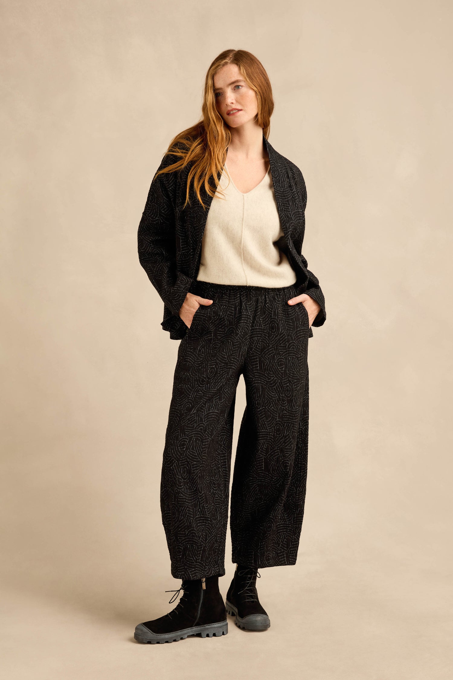 A person stands with hands in pockets, wearing a textured black jacket, Stitched Jacquard Bubble Trouser with a cropped leg, a cream V-neck top, and black boots against a beige background.