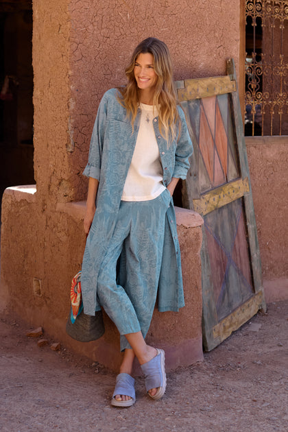 Woman in blue Floral Jacquard Cotton Bubble Trouser set and sandals leans against a rustic clay wall, smiling, with a colorful bag and wooden door in the background.