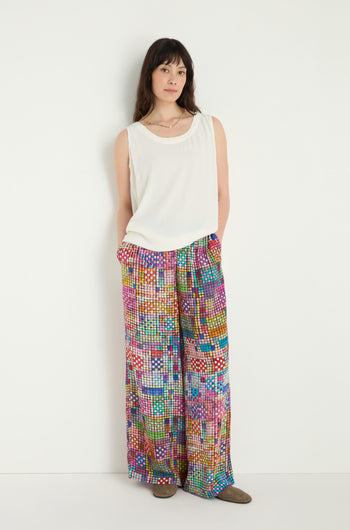 A woman stands against a white wall wearing a sleeveless white top, flat brown shoes, and Coloured Glass Tiles Wide Trousers with vibrant patterns, her hands casually in her pockets.