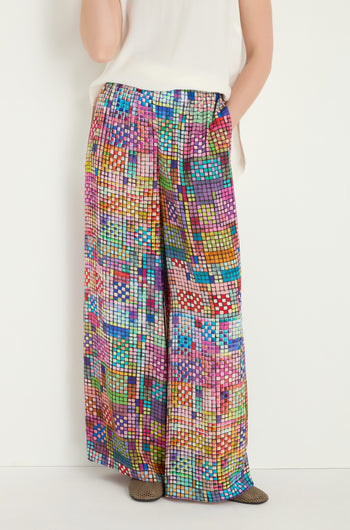 Person wearing Coloured Glass Tiles Wide Trousers—loose, wide-leg pants with a pixel-like pattern—and a white sleeveless top, hands in pockets, standing against a plain background.