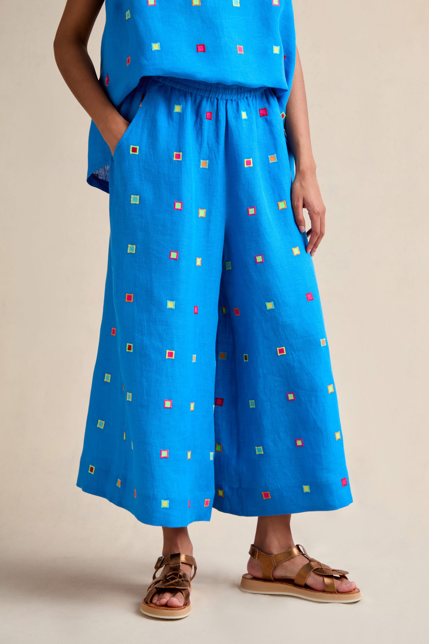 Wearing the Embroidered Squares Linen Wide Trousers in bright blue with a matching cropped top and brown sandals, this person stands against a neutral background.