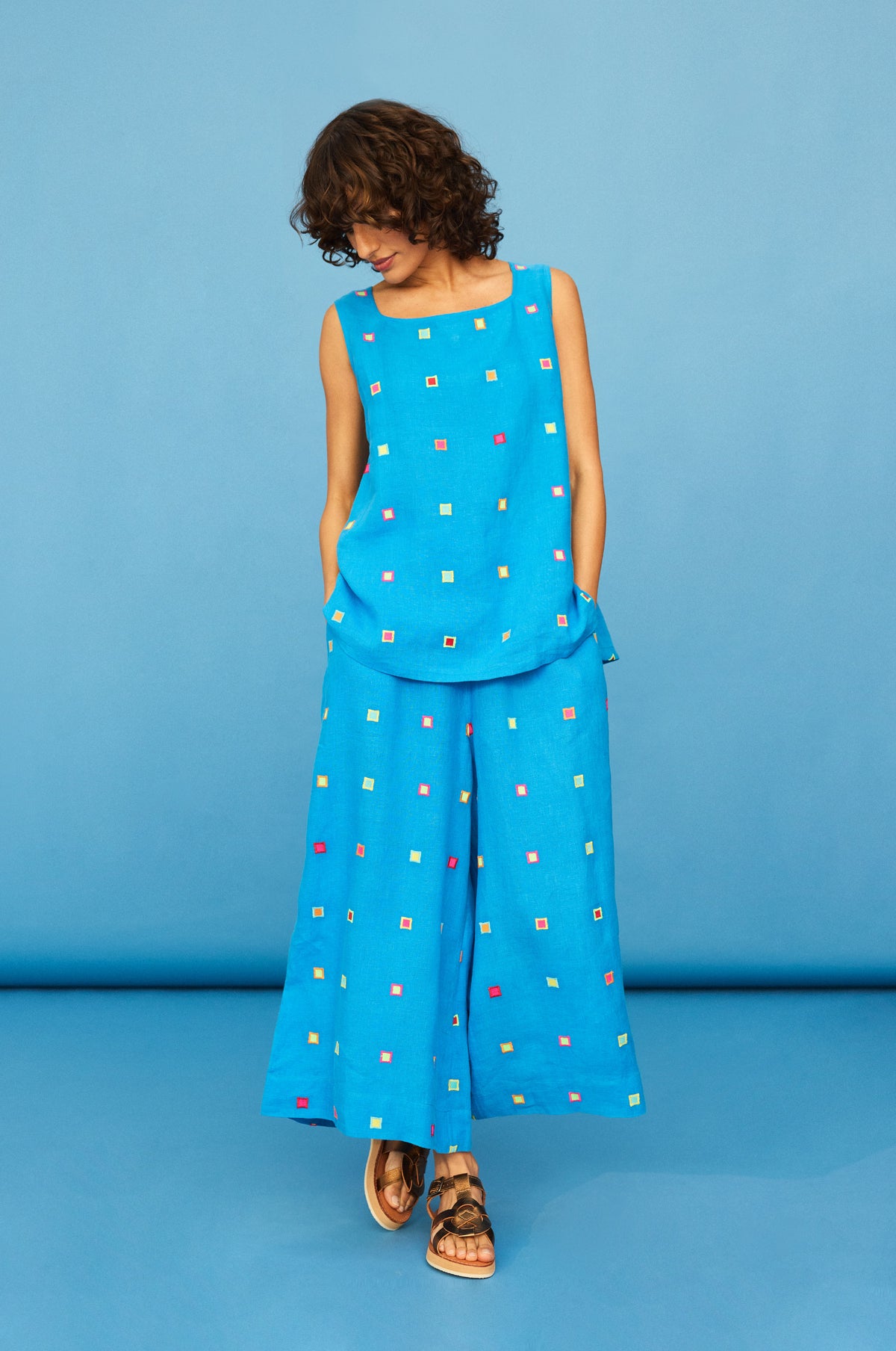Someone stands against a blue background, hands in pockets, wearing Embroidered Squares Linen Wide Trousers in a cropped length with an embroidered squares pattern and a sleeveless blue top, looking down.