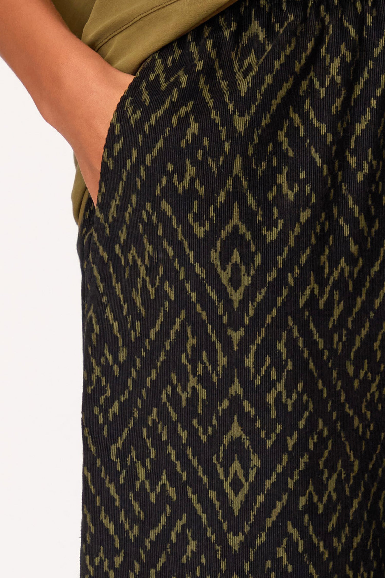 A close-up of a hand slipped into the pocket of Ornate Textured Cord Bubble Trousers in black and olive green, featuring a sculptural silhouette and styled with an olive green top.