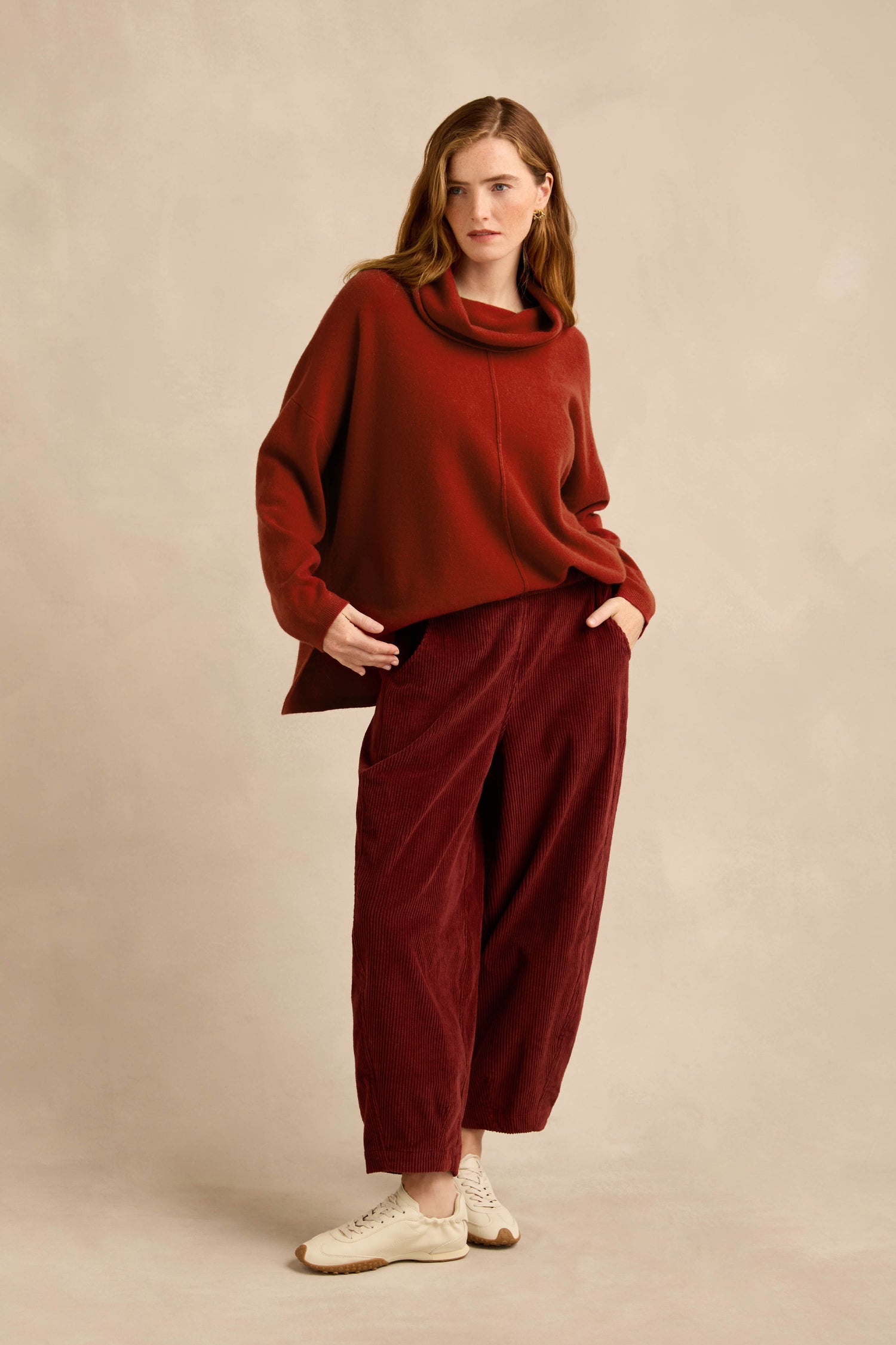 A woman stands against a beige background with her hands in her pockets, wearing a loose red sweater, the Conker Jumbo Cord Bubble Trouser in a wide red silhouette with a comfortable pull-on style, and white sneakers.