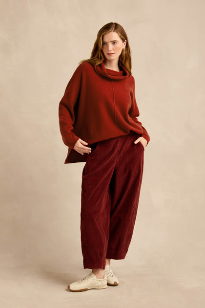 A woman stands against a beige background with her hands in her pockets, wearing a loose red sweater, the Conker Jumbo Cord Bubble Trouser in a wide red silhouette with a comfortable pull-on style, and white sneakers.
