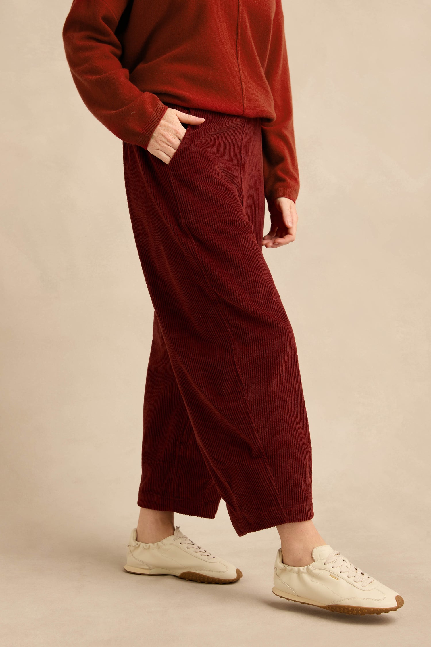 A person stands with one hand in their pocket, wearing the Conker Jumbo Cord Bubble Trouser—a rust-red, pull-on cotton cord style—paired with a matching sweater and white sneakers.