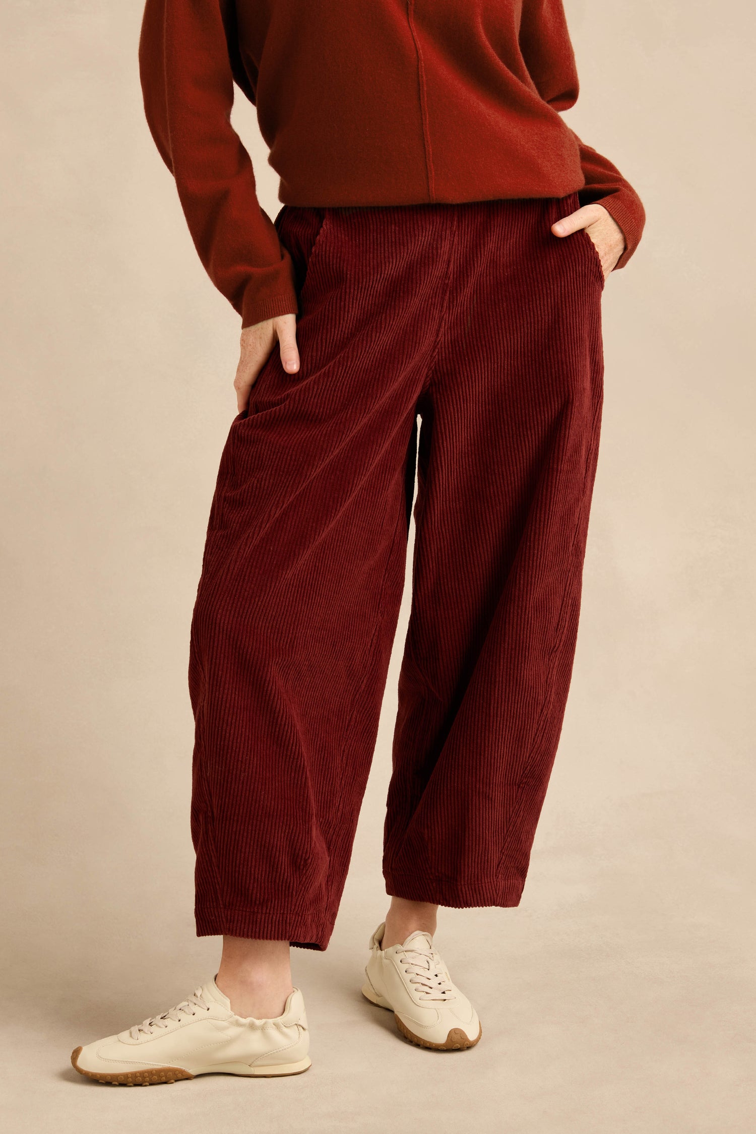 A person wears the Conker Jumbo Cord Bubble Trouser in burgundy—a cotton cord pull-on style—paired with a long-sleeve top and white sneakers, standing with one hand in their pocket.