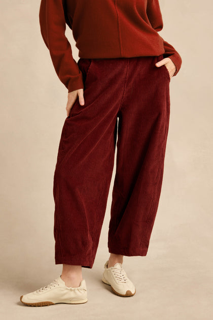 A person wears the Conker Jumbo Cord Bubble Trouser in burgundy—a cotton cord pull-on style—paired with a long-sleeve top and white sneakers, standing with one hand in their pocket.