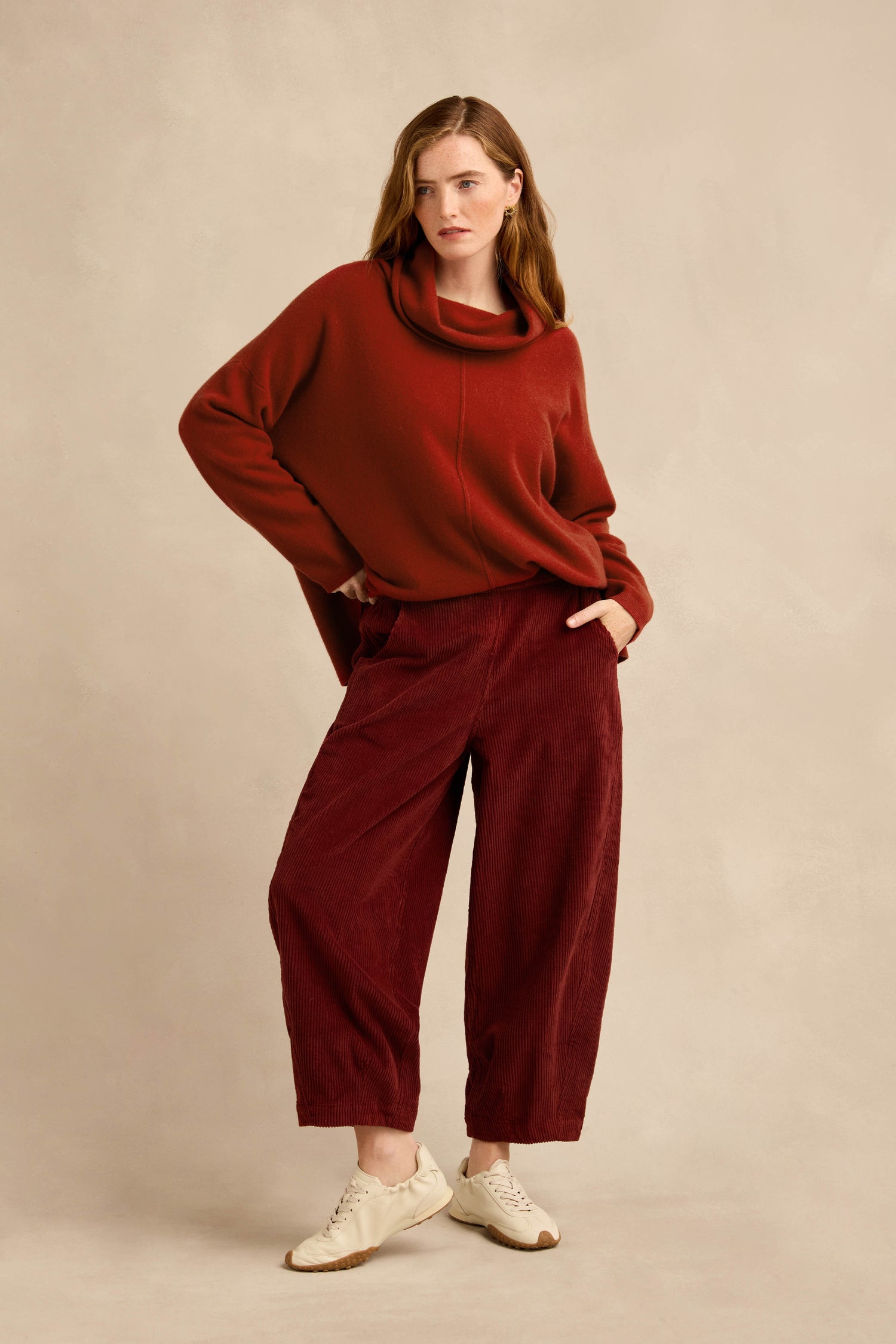 A woman stands against a neutral background wearing a loose red sweater with a bubble silhouette, the Conker Jumbo Cord Bubble Trouser in maroon with wide legs and an easy pull-on style, and white sneakers.