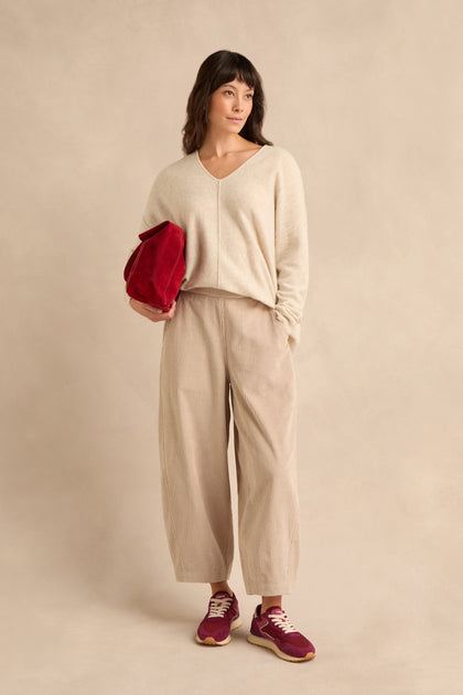 Certainly! Here’s a rewritten version using the provided product name: A woman stands against a beige background, wearing a beige sweater and Ecru Jumbo Cord Bubble Trousers with a wide-leg, bubble silhouette, paired with red sneakers and holding a red bag.