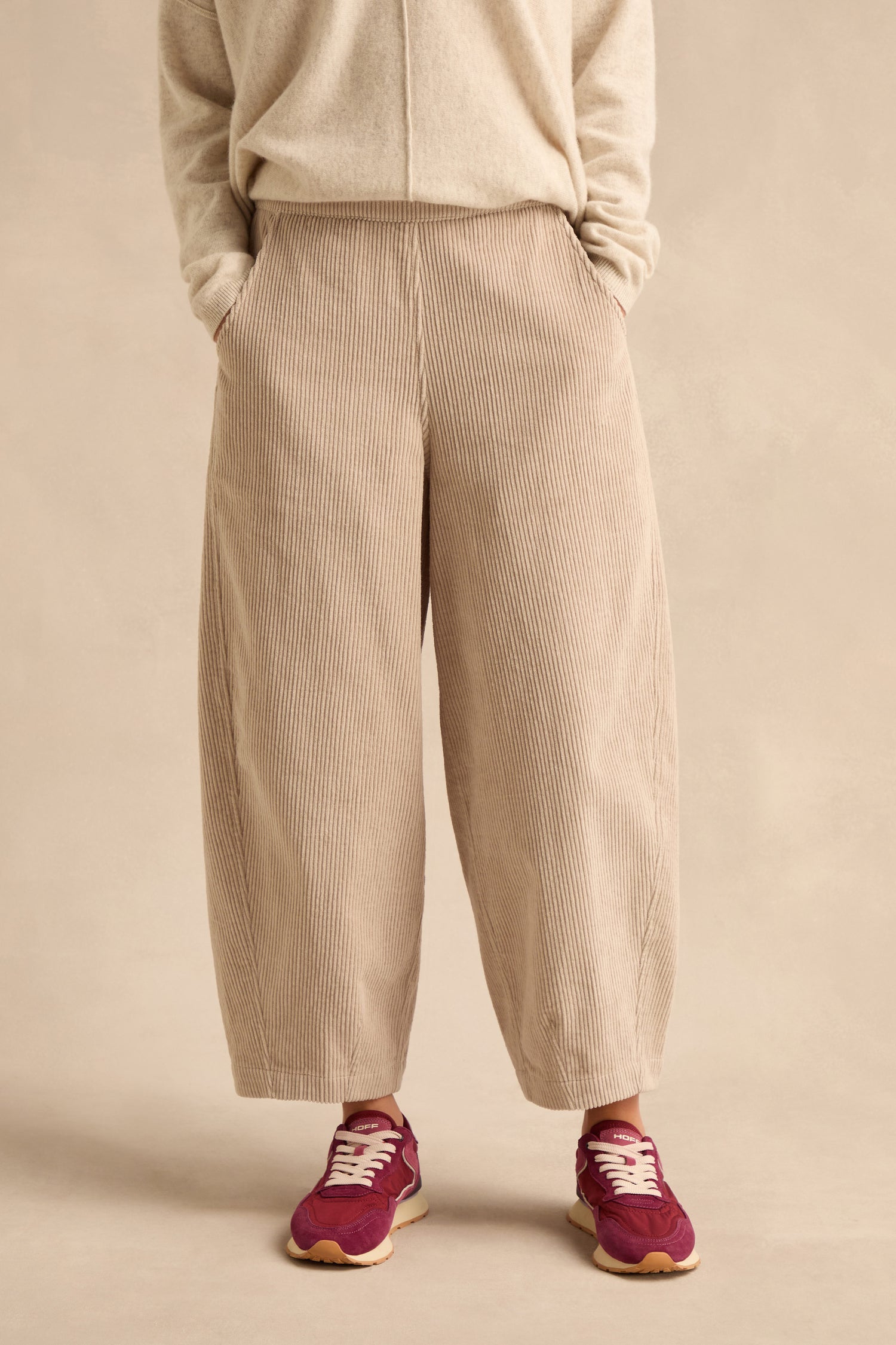 Wearing the Ecru Jumbo Cord Bubble Trousers in a wide-leg, bubble silhouette, paired with a matching sweater and red sneakers, this person stands with hands in pockets against a neutral background.