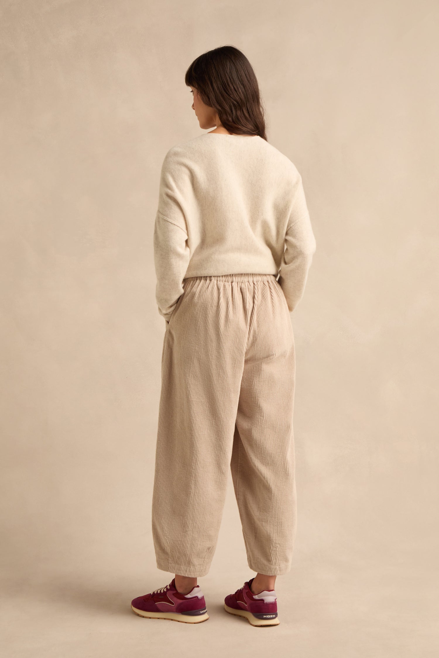 A person stands with their back to the camera, wearing a cream sweater and the Ecru Jumbo Cord Bubble Trousers in a relaxed bubble silhouette, paired with maroon sneakers, set against a neutral beige background.