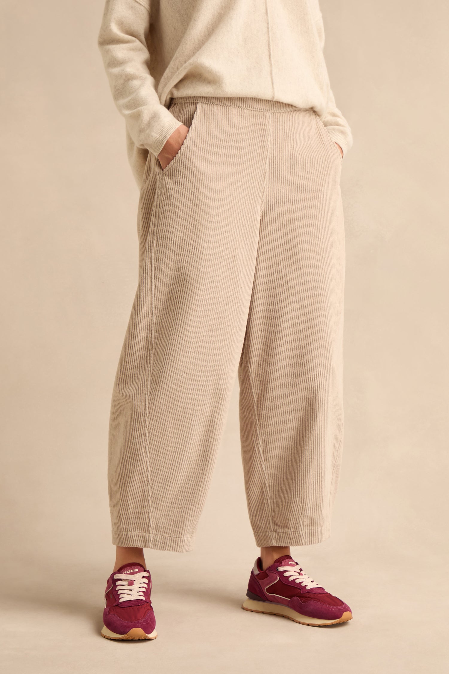 A person stands with hands in pockets against a plain background, wearing Ecru Jumbo Cord Bubble Trousers in a beige shade, paired with a matching sweater and purple sneakers.