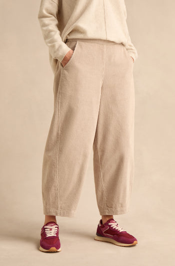 A person stands with hands in pockets against a plain background, wearing Ecru Jumbo Cord Bubble Trousers in a beige shade, paired with a matching sweater and purple sneakers.