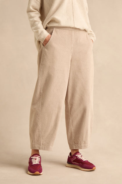 A person stands with hands in pockets against a plain background, wearing Ecru Jumbo Cord Bubble Trousers in a beige shade, paired with a matching sweater and purple sneakers.