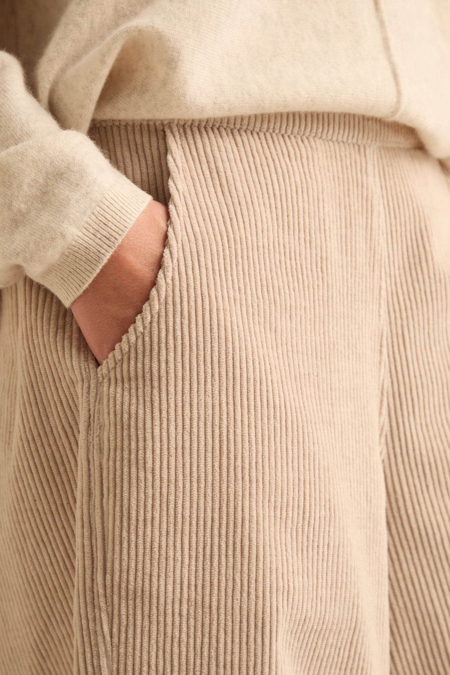 Close-up of a person’s hand in the pocket of Ecru Jumbo Cord Bubble Trousers, paired with a matching beige sweater.
