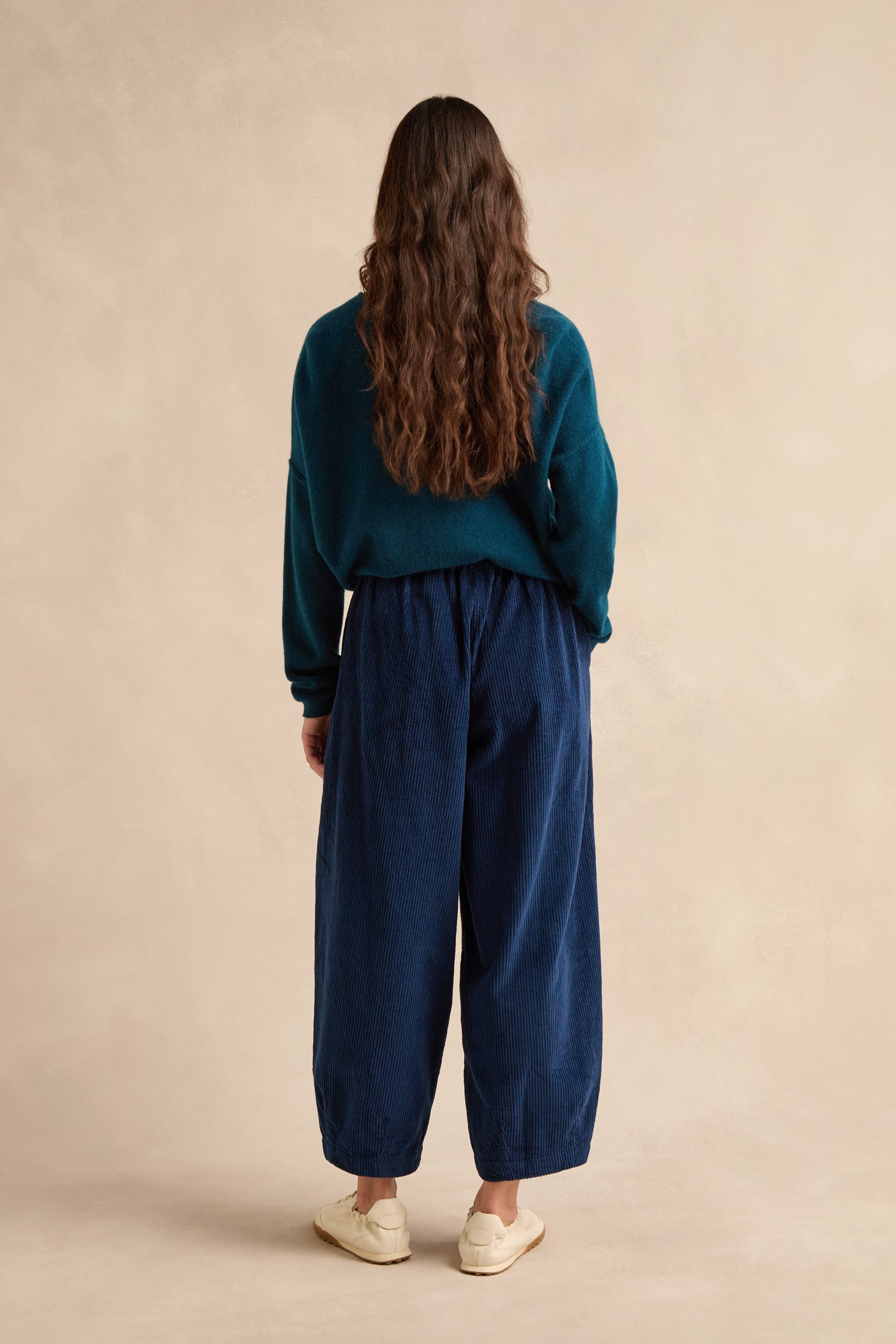 A person with long brown hair, wearing a teal sweater, Marine Jumbo Cord Bubble Trousers in navy with a bubble silhouette, and beige shoes stands facing away from the camera against a neutral background.