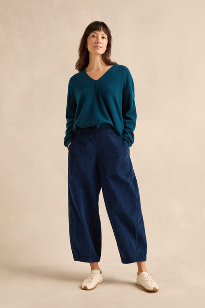 A woman stands against a plain beige background, wearing a teal V-neck sweater paired with the Marine Jumbo Cord Bubble Trousers in dark blue, styled with white sneakers and her hands tucked into her pockets.