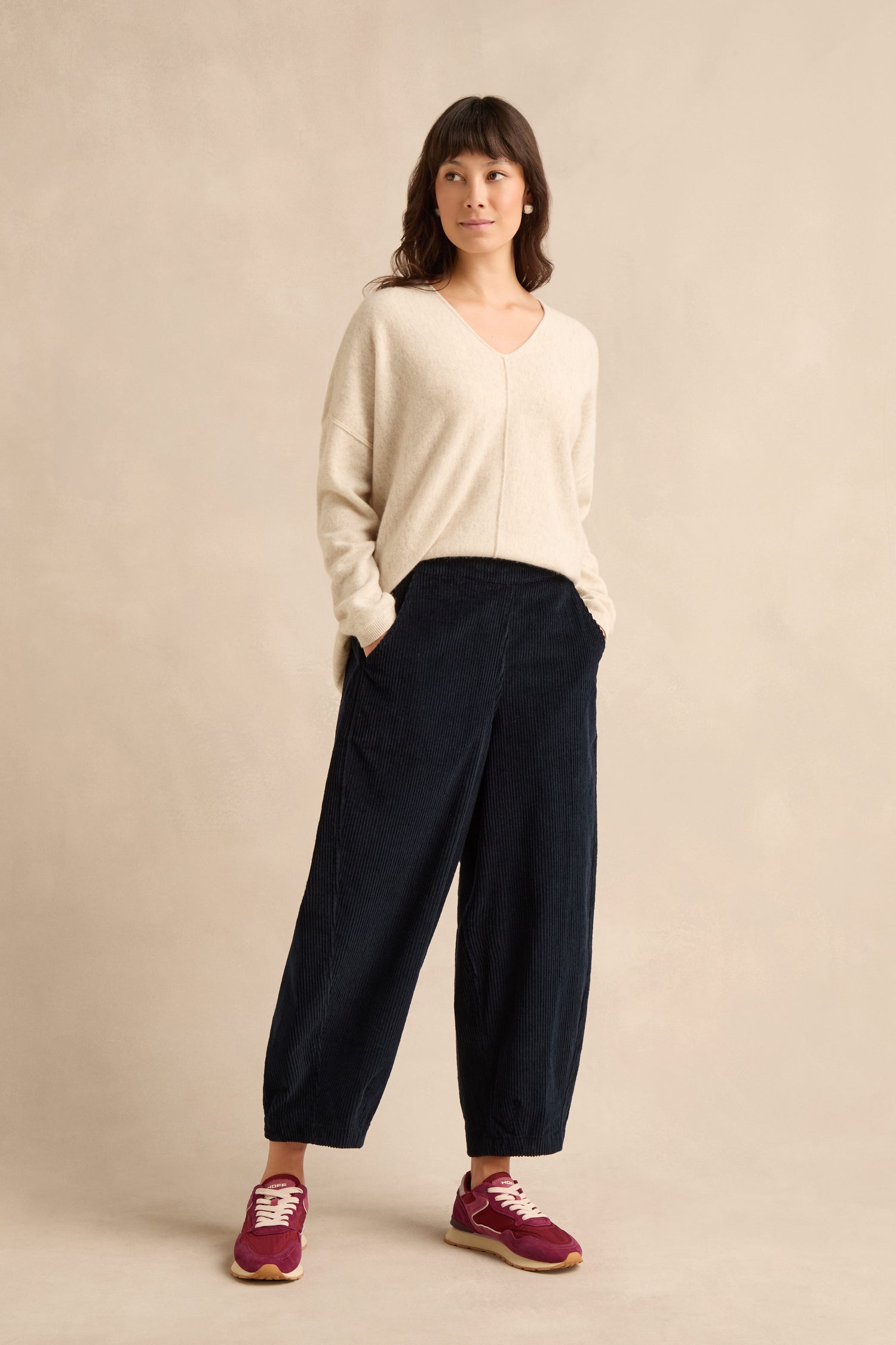 Woman standing against a beige background, wearing a cream pure cotton sweater, Navy Jumbo Cord Bubble Trousers with a distinctive bubble silhouette, and red sneakers, with hands in pockets.