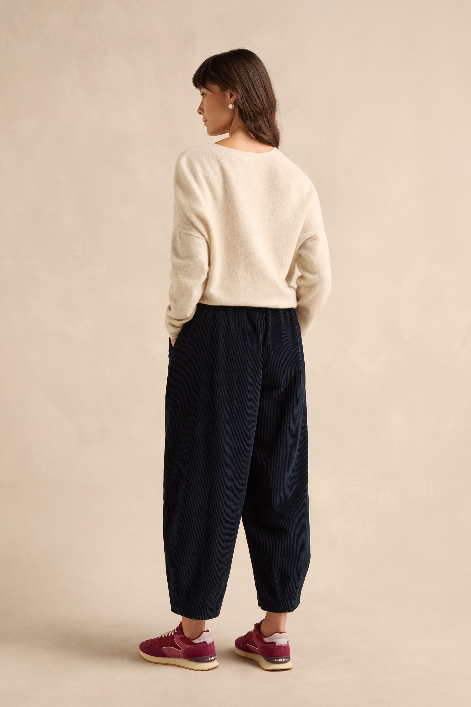 A person stands facing away, wearing a cream sweater, pure cotton Navy Jumbo Cord Bubble Trousers with a bubble silhouette, and red sneakers, against a plain beige background.
