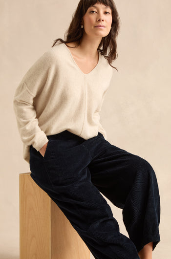 A woman in a cream pure cotton V-neck sweater and navy jumbo cord bubble trousers sits on a wooden block against a plain beige background.