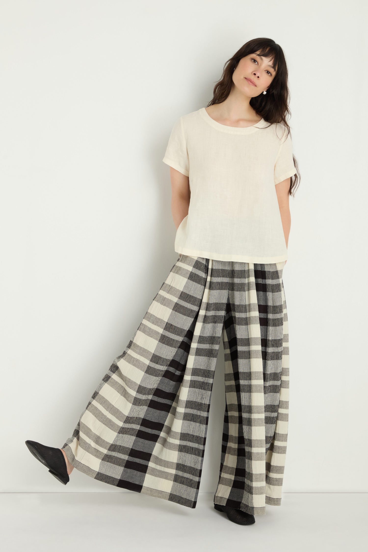 A woman stands against a plain background, one foot slightly raised, wearing a short-sleeved beige top and Monochrome Crinkle Check Wide Trouser, hands in her pockets.