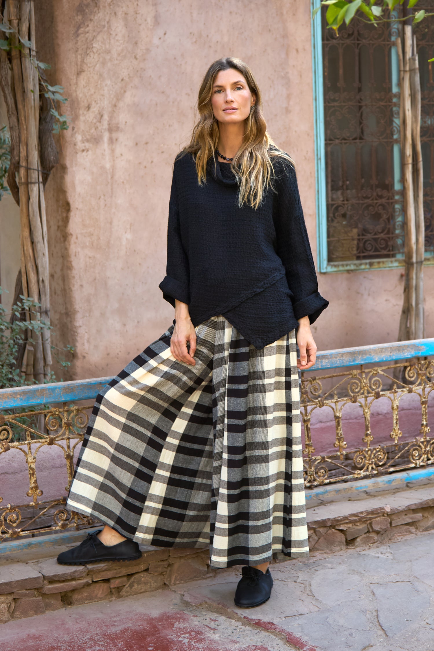 A woman stands outdoors beside a rustic metal railing, wearing Monochrome Crinkle Check Wide Trousers and a black long-sleeve top, with plants and a weathered wall in the background.