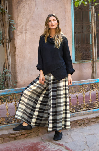 A woman stands outdoors beside a rustic metal railing, wearing Monochrome Crinkle Check Wide Trousers and a black long-sleeve top, with plants and a weathered wall in the background.
