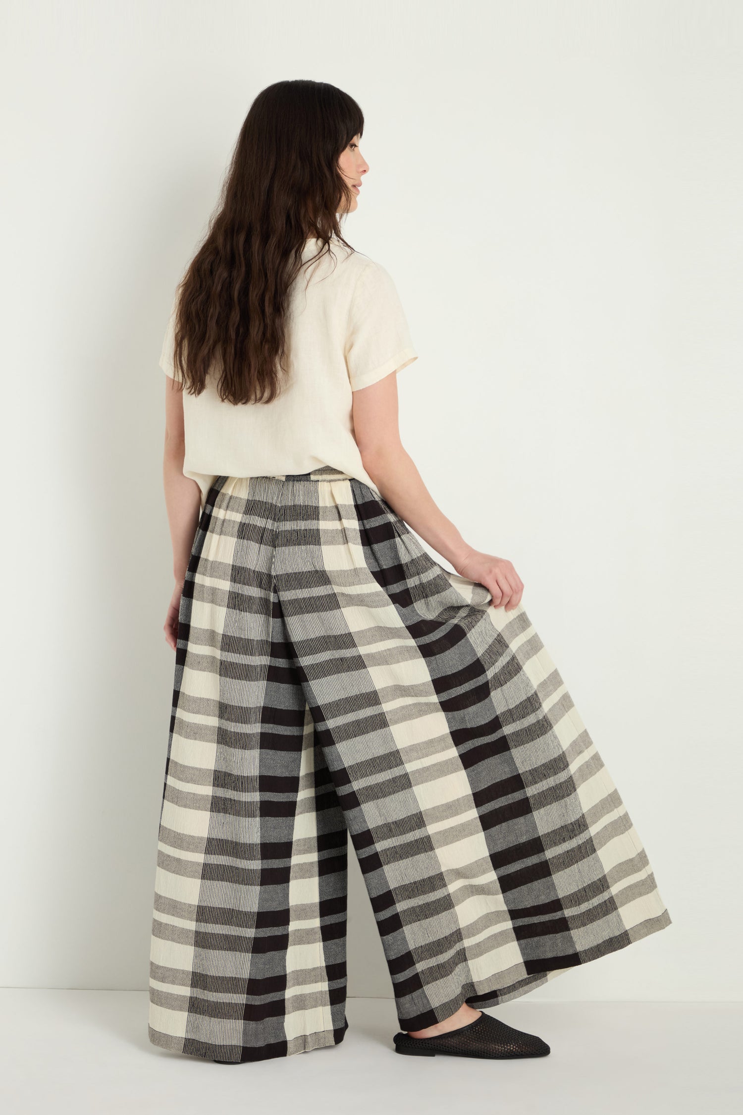 A person stands facing away, wearing a cream short-sleeve top and the Monochrome Crinkle Check Wide Trouser—travel-friendly, wide-leg pants in a black and white check pattern—paired with black flats against a plain background.