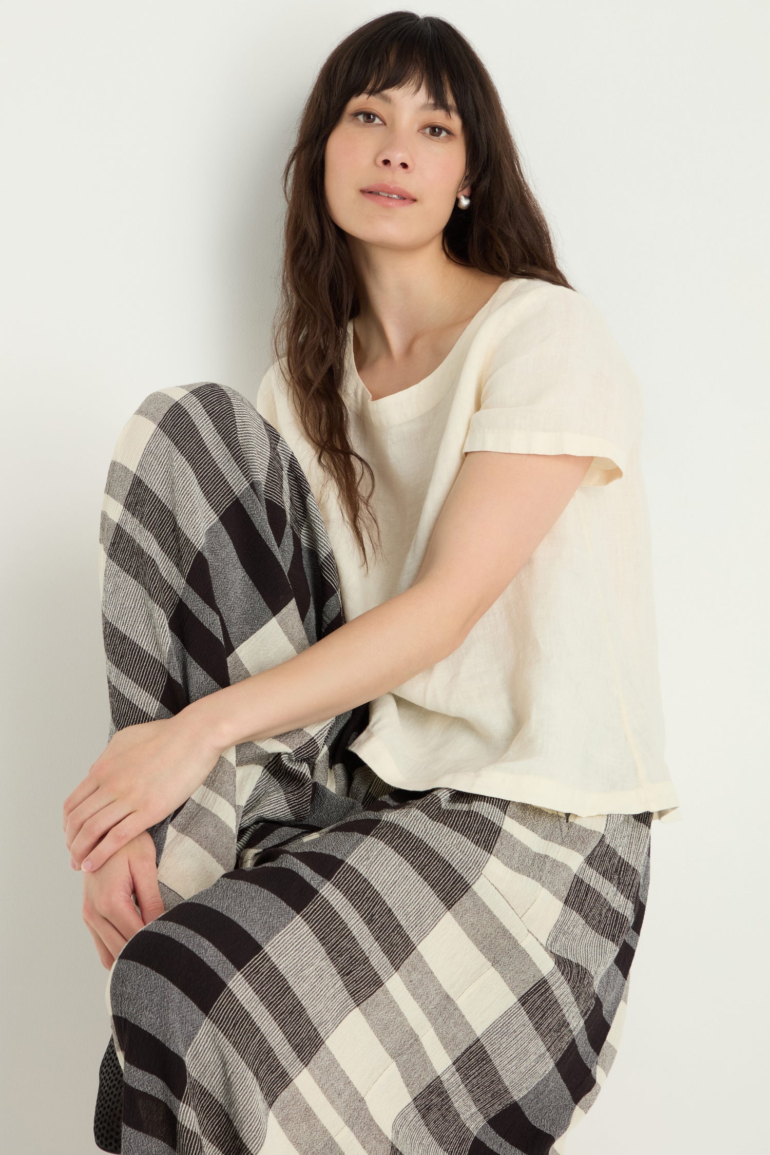 A woman sits with one knee raised, wearing a short-sleeve cream top and Monochrome Crinkle Check Wide Trousers against a plain light background.