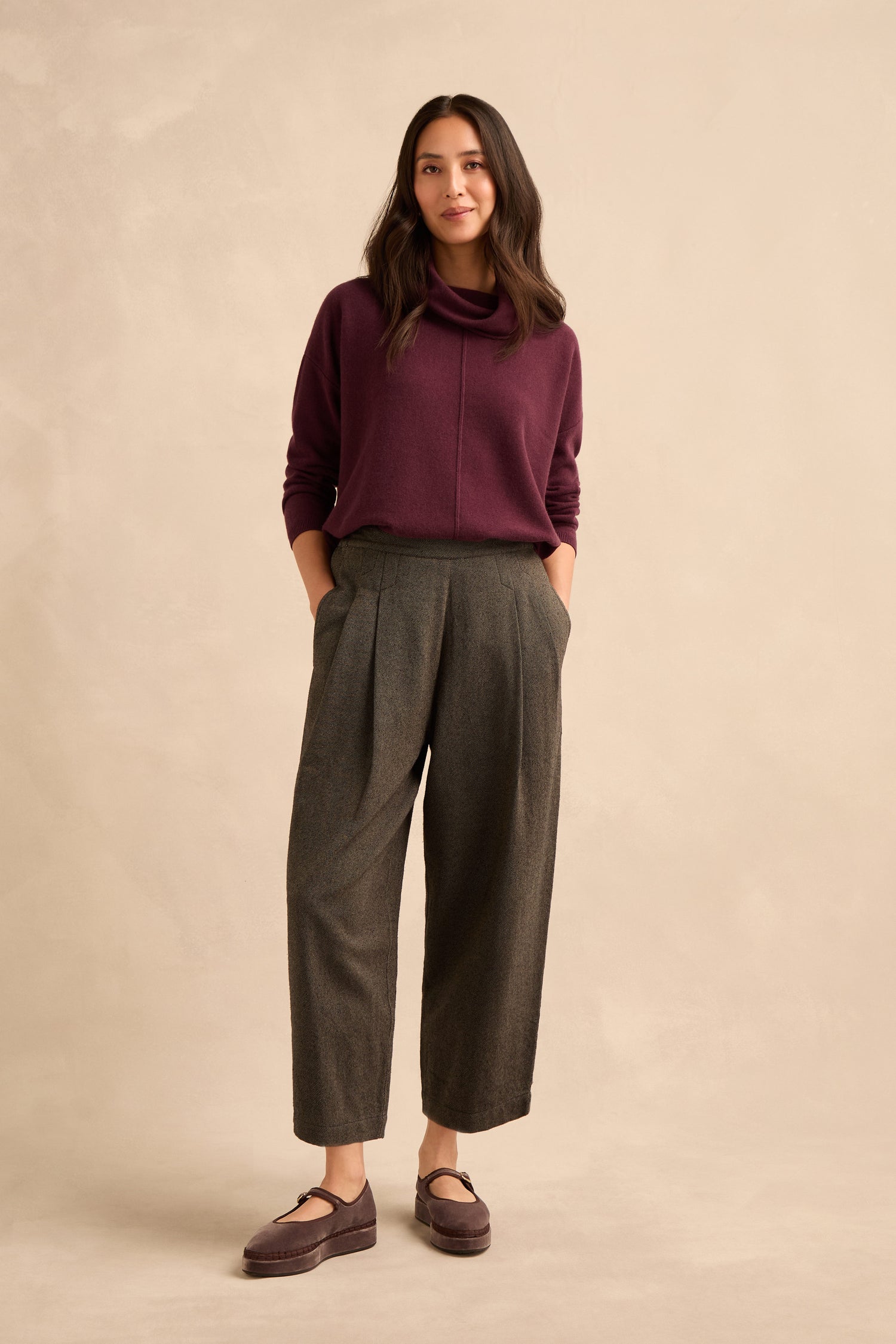 A woman stands against a beige background, wearing a maroon sweater, Overdyed Twill Barrel Trouser in gray cotton, and mauve loafers, with her hands in her pockets.