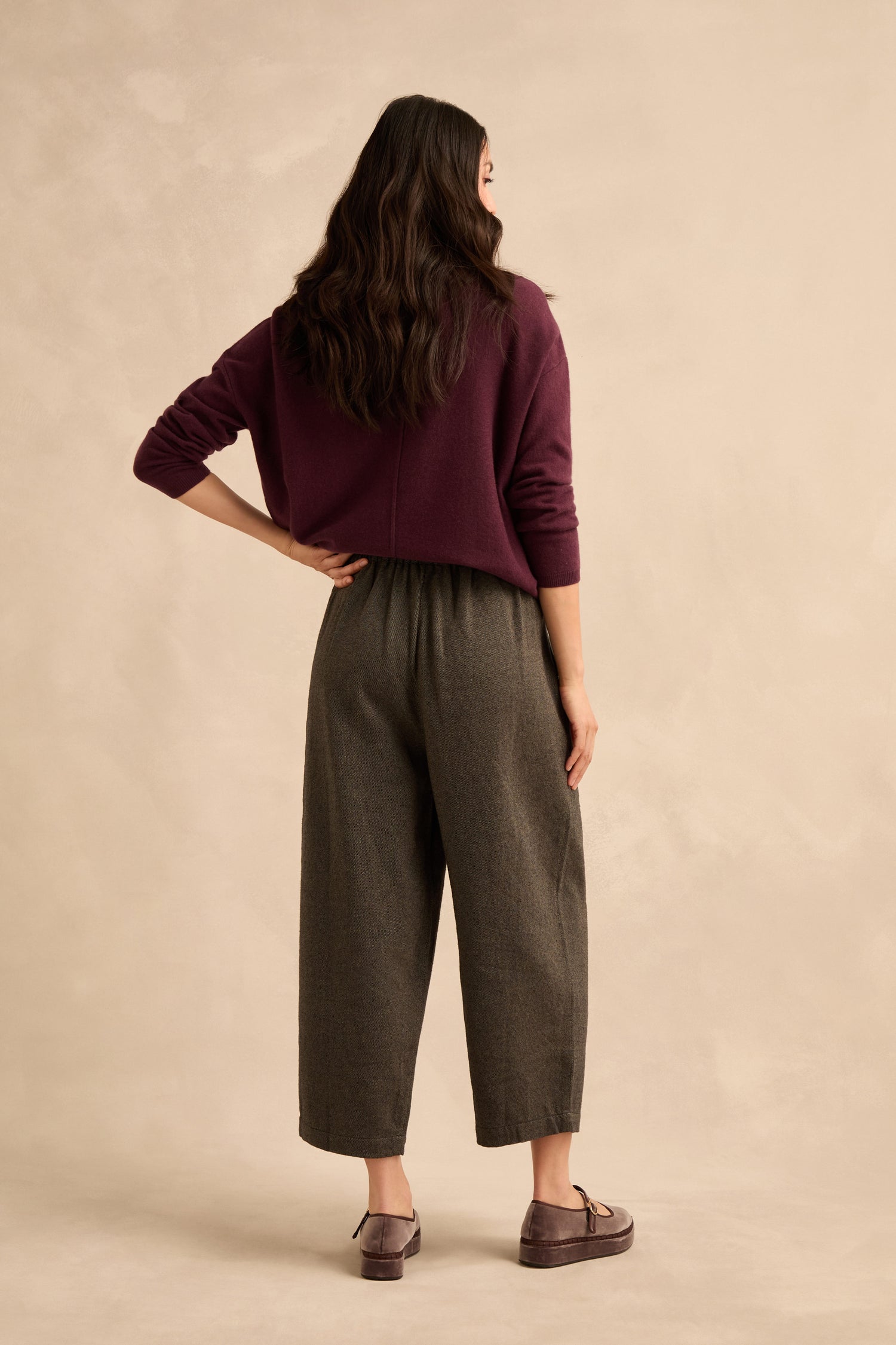 A woman stands facing away, wearing a maroon sweater, Overdyed Twill Barrel Trousers in gray, and brown shoes, against a plain beige background.