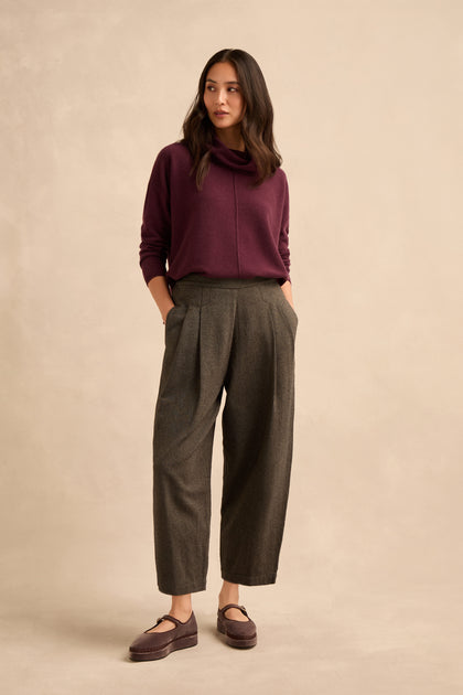 A woman stands against a neutral background, wearing a burgundy sweater and the Overdyed Twill Barrel Trouser in loose gray pleats, paired with mauve shoes. She has her hands in her pockets and looks slightly to the side.