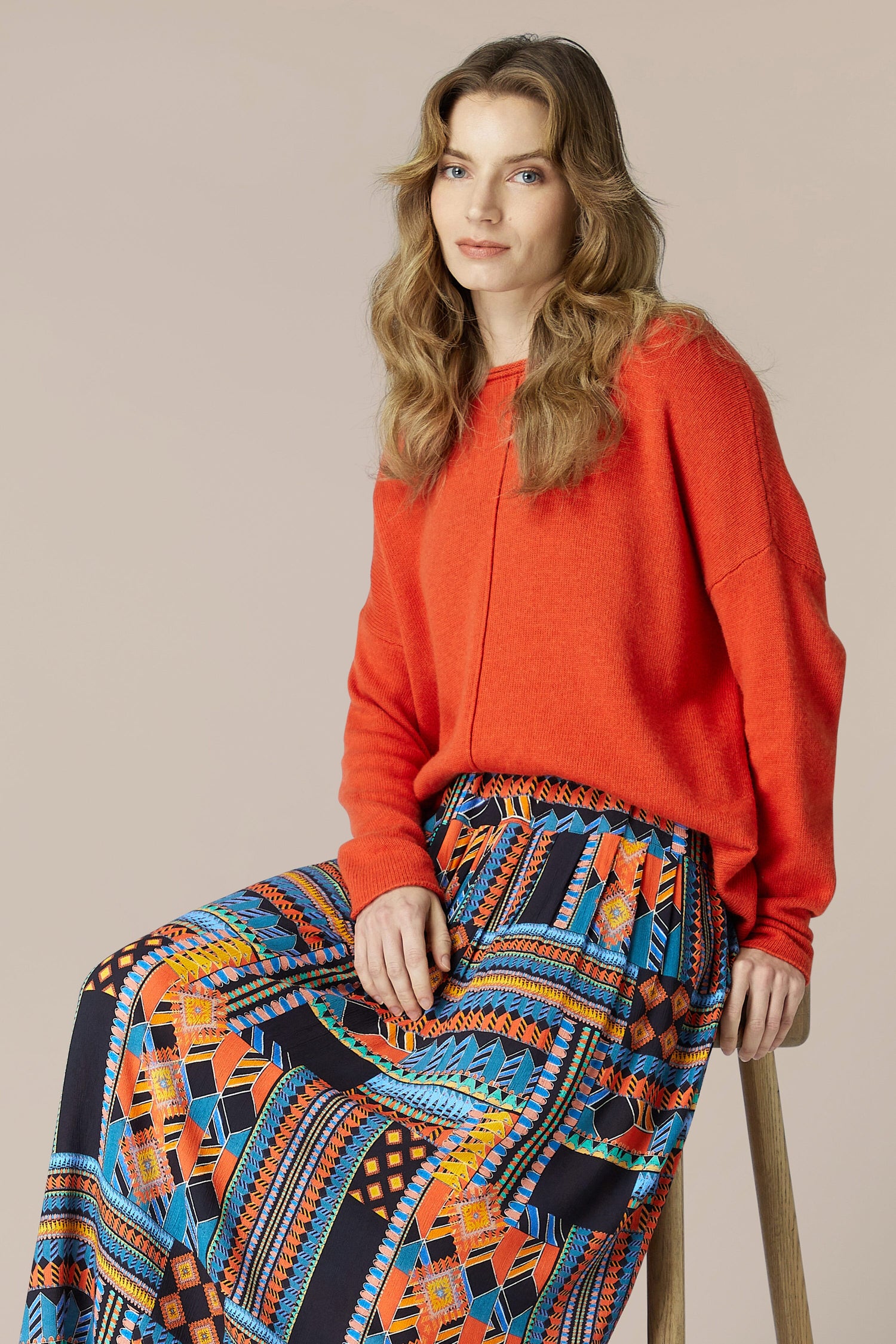 A woman is sitting on a stool wearing a vibrant orange sweater and a flowing midi-length Bold Graphic Printed Skirt made of viscose.