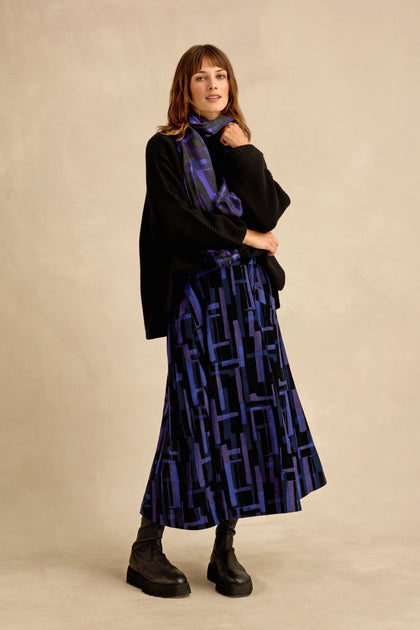A woman wearing a black knit sweater, the Layered Geometric Cord Skirt in blue and purple with a matching scarf, and black platform boots stands against a beige background.
