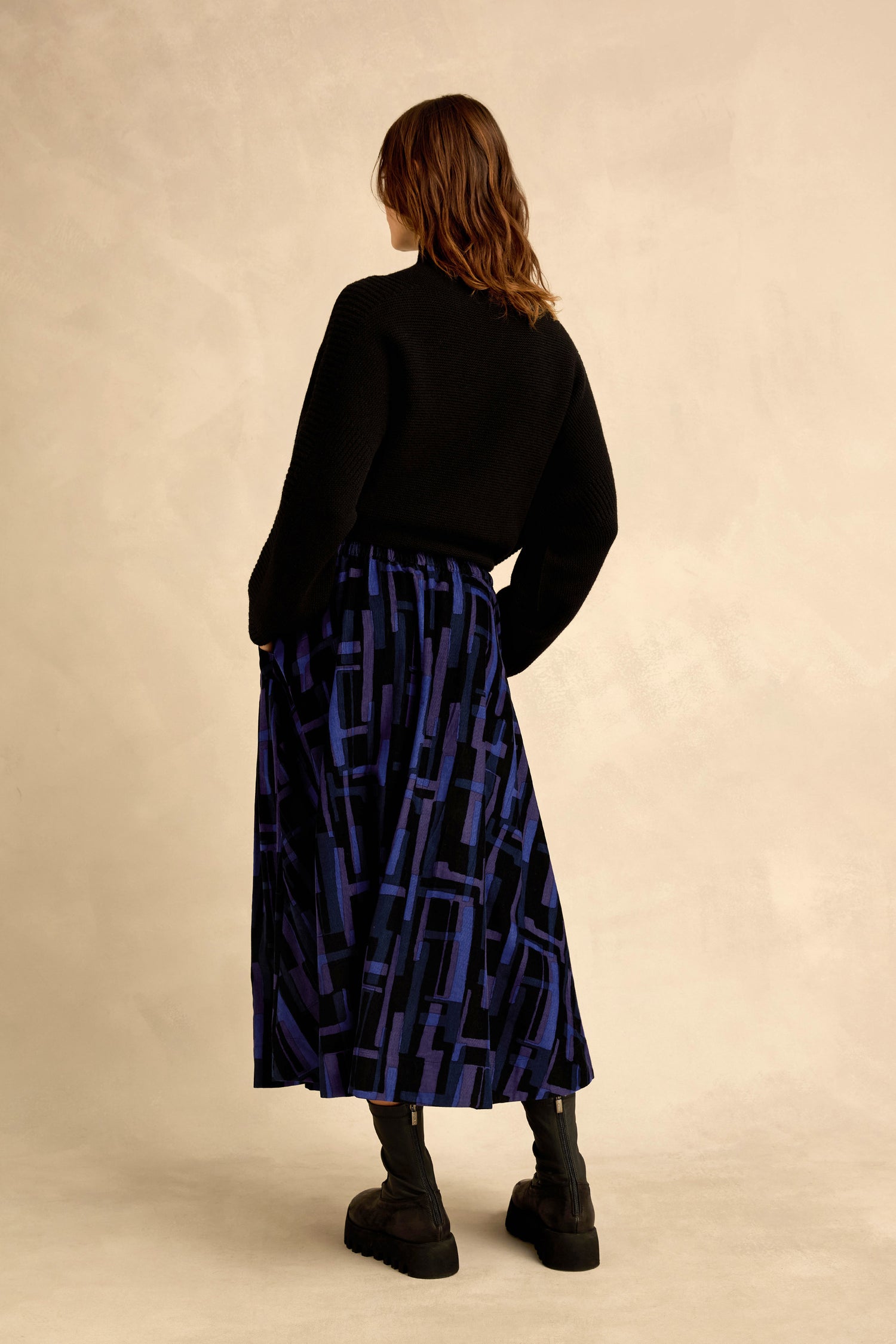 Person standing with their back to the camera, wearing a black sweater, the Layered Geometric Cord Skirt featuring a long blue and black geometric pattern, and black boots against a neutral background.