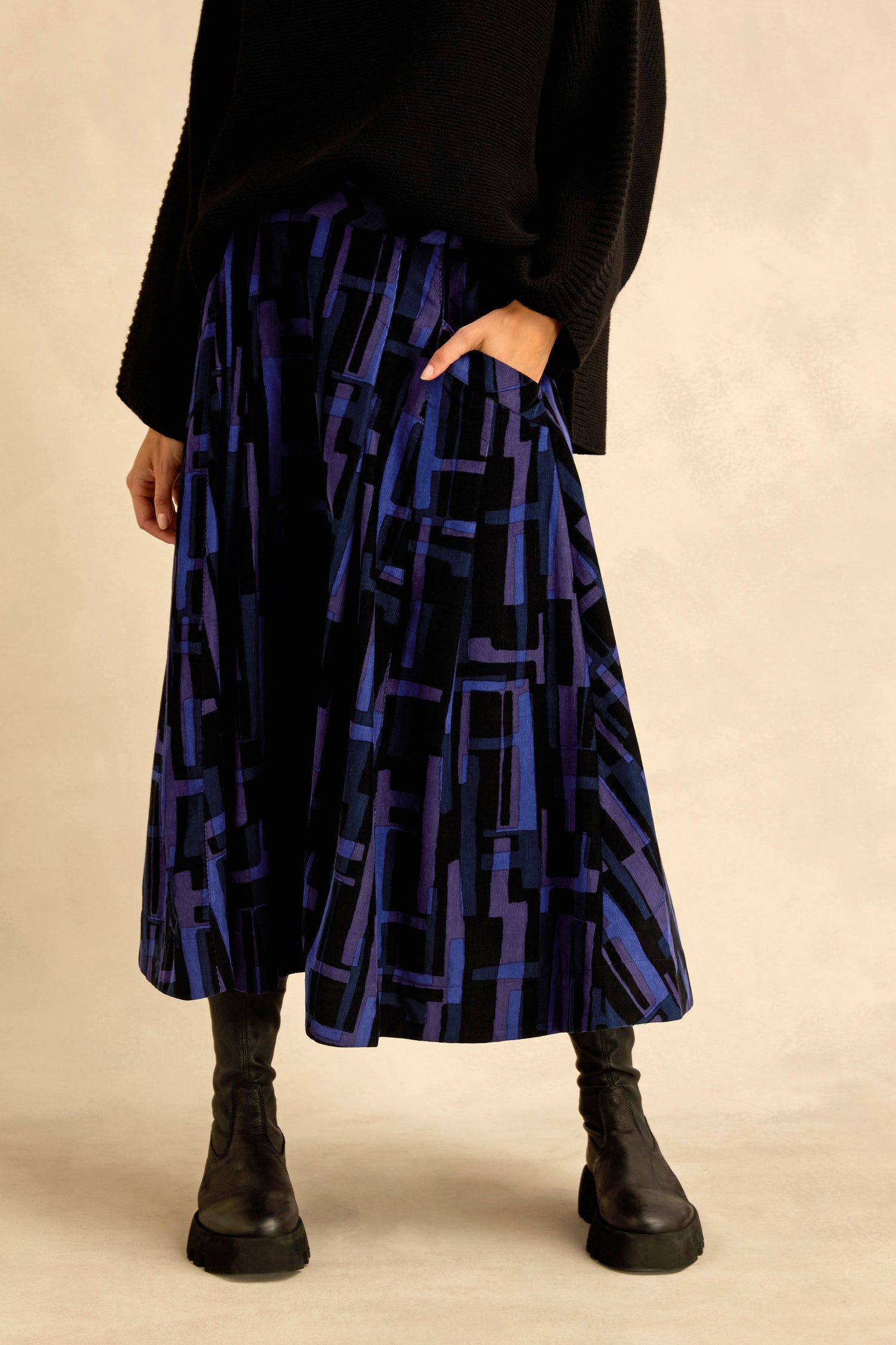 Person wearing a black sweater, the Layered Geometric Cord Skirt in blue and black cotton with a geometric pattern, and black boots, standing with one hand in their pocket against a neutral background.