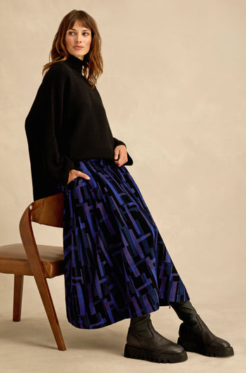 A woman in a black sweater and black boots sits sideways on a wooden chair against a beige background, highlighting the bold allure of her Layered Geometric Cord Skirt.