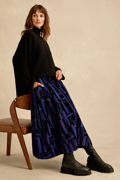 A woman in a black sweater and black boots sits sideways on a wooden chair against a beige background, highlighting the bold allure of her Layered Geometric Cord Skirt.