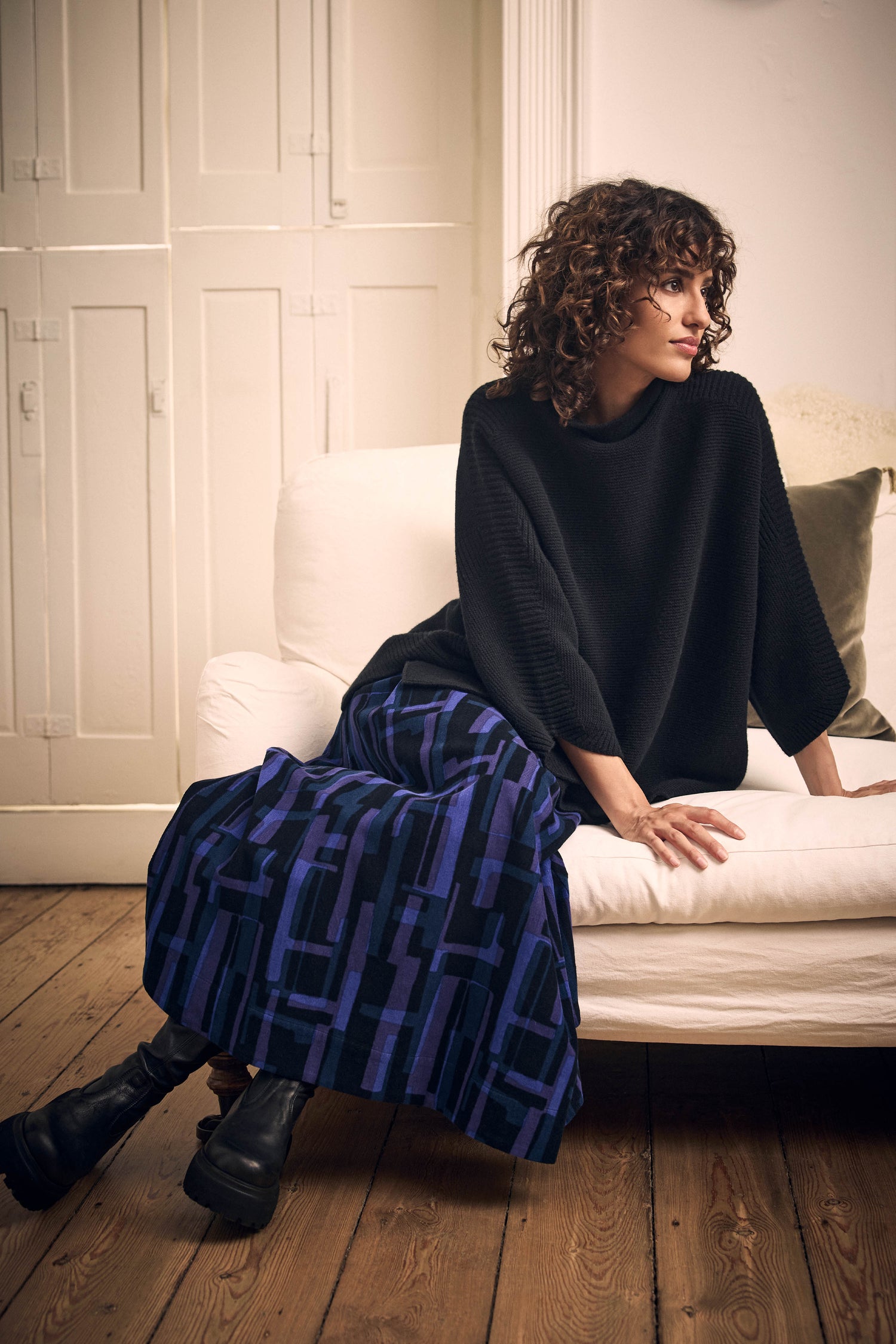 A person with curly hair wearing a black sweater and a Layered Geometric Fine Cord Skirt with a blue and purple pattern, sitting on a white sofa.