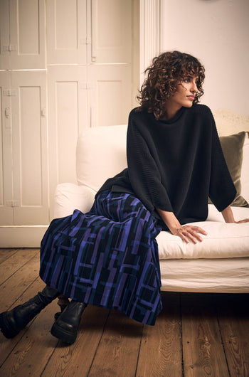 A person with curly hair wearing a black sweater and a Layered Geometric Fine Cord Skirt with a blue and purple pattern, sitting on a white sofa.