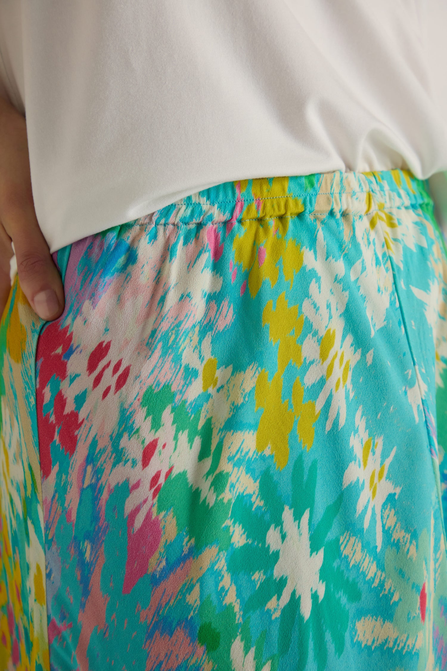 Close-up of a person wearing the Floral Oasis Bias Skirt, featuring a vibrant floral print and an elastic waistband, paired with a white top. Their left hand rests on their hip.