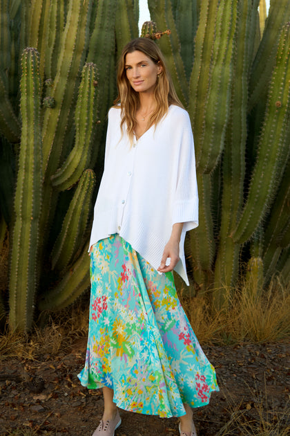 A woman stands before large cacti, wearing a white buttoned sweater, tan shoes, and the Floral Oasis Bias Skirt.
