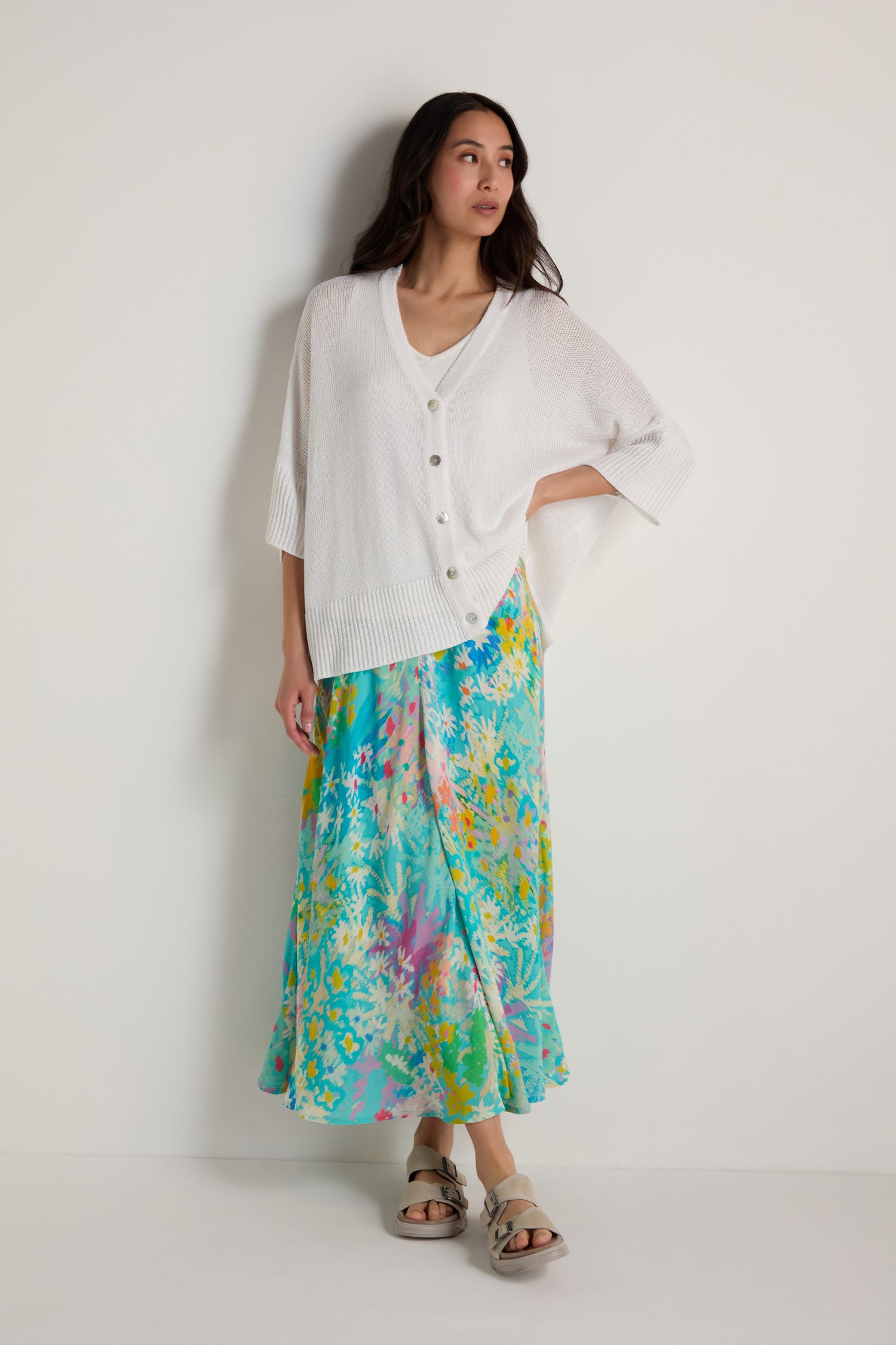 A woman stands against a plain white background wearing a loose white cardigan over a cream top, paired with the Floral Oasis Bias Skirt and beige sandals.