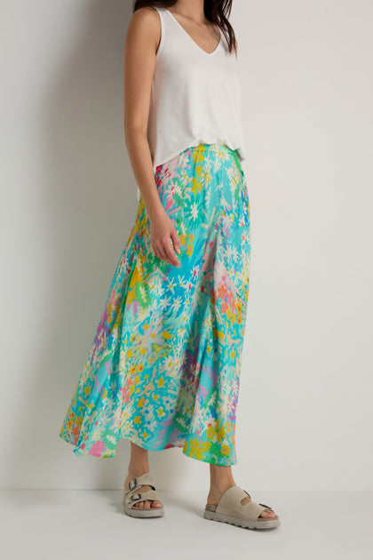 A woman stands against a white wall wearing a white sleeveless top, the Floral Oasis Bias Skirt in blue floral, and beige sandals.