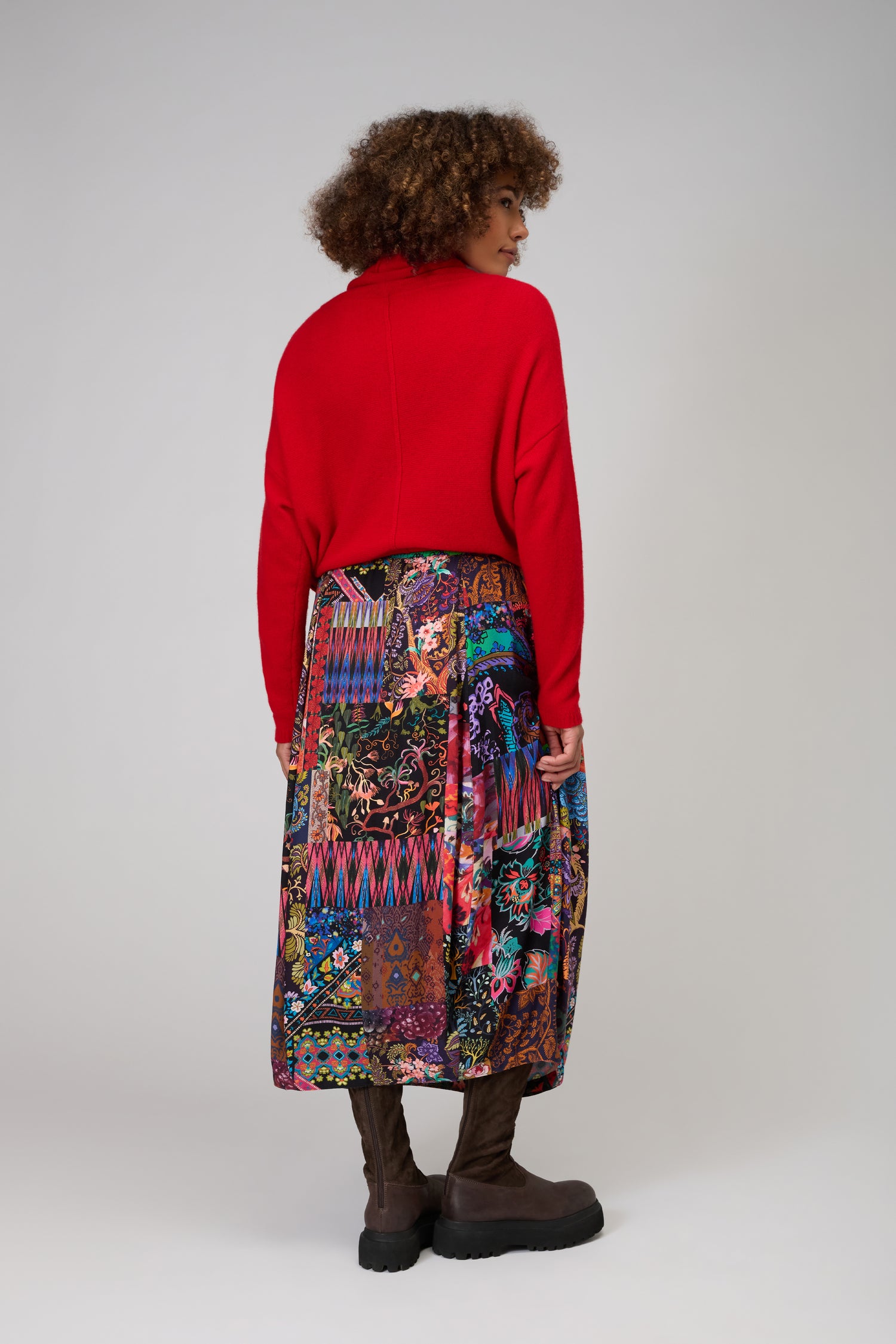 A person with curly hair, in a red sweater, Sahara Winter Patchwork Skirt, and brown boots stands facing away against a neutral background.