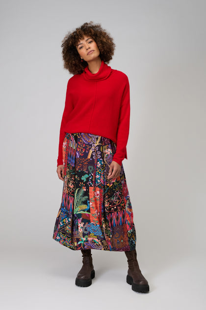A person stands against a plain light gray background, wearing a red long-sleeved top, dark brown boots, and the Sahara Winter Patchwork Skirt with a bubble silhouette.