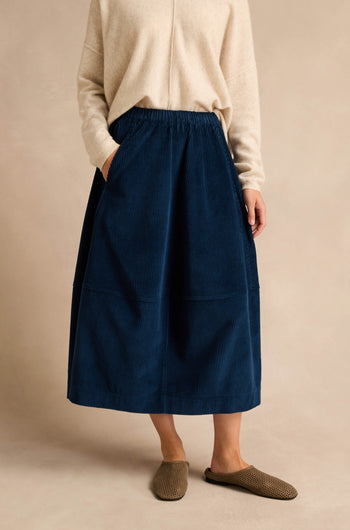 Person wearing a cream long-sleeve top, the Jumbo Cord Bubble Skirt in blue with pockets, and brown slip-on shoes, standing against a neutral background.